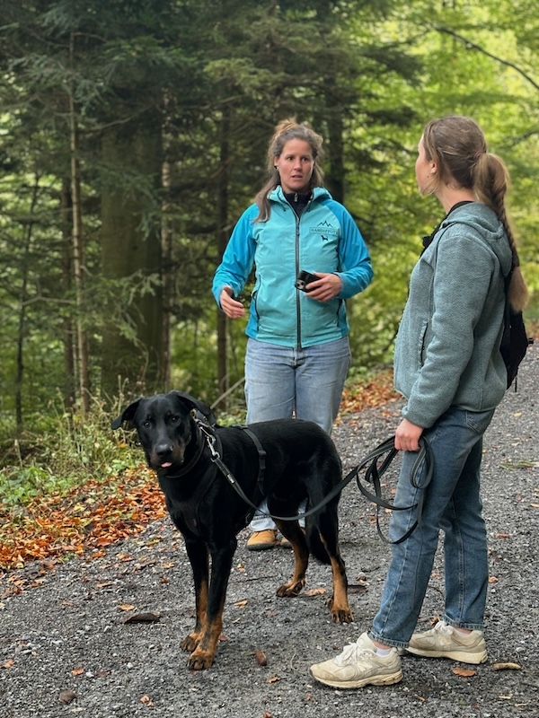 Einzeltraining im Wald: Linda erklärt einer Frau etwas, die Frau schaut Linda an. Ihr Hund steht neben ihr an der Leine und schaut in die Kamera