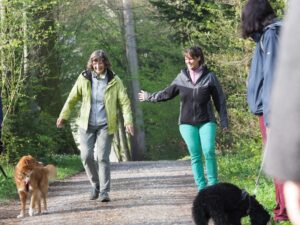 Hundetraining im Wald: Linda und eine Frau laufen an einem Hund vorbei, der an der Leine läuft und sie anschaut. Vorne wartet ein anderer Hund mit seiner Besitzerin